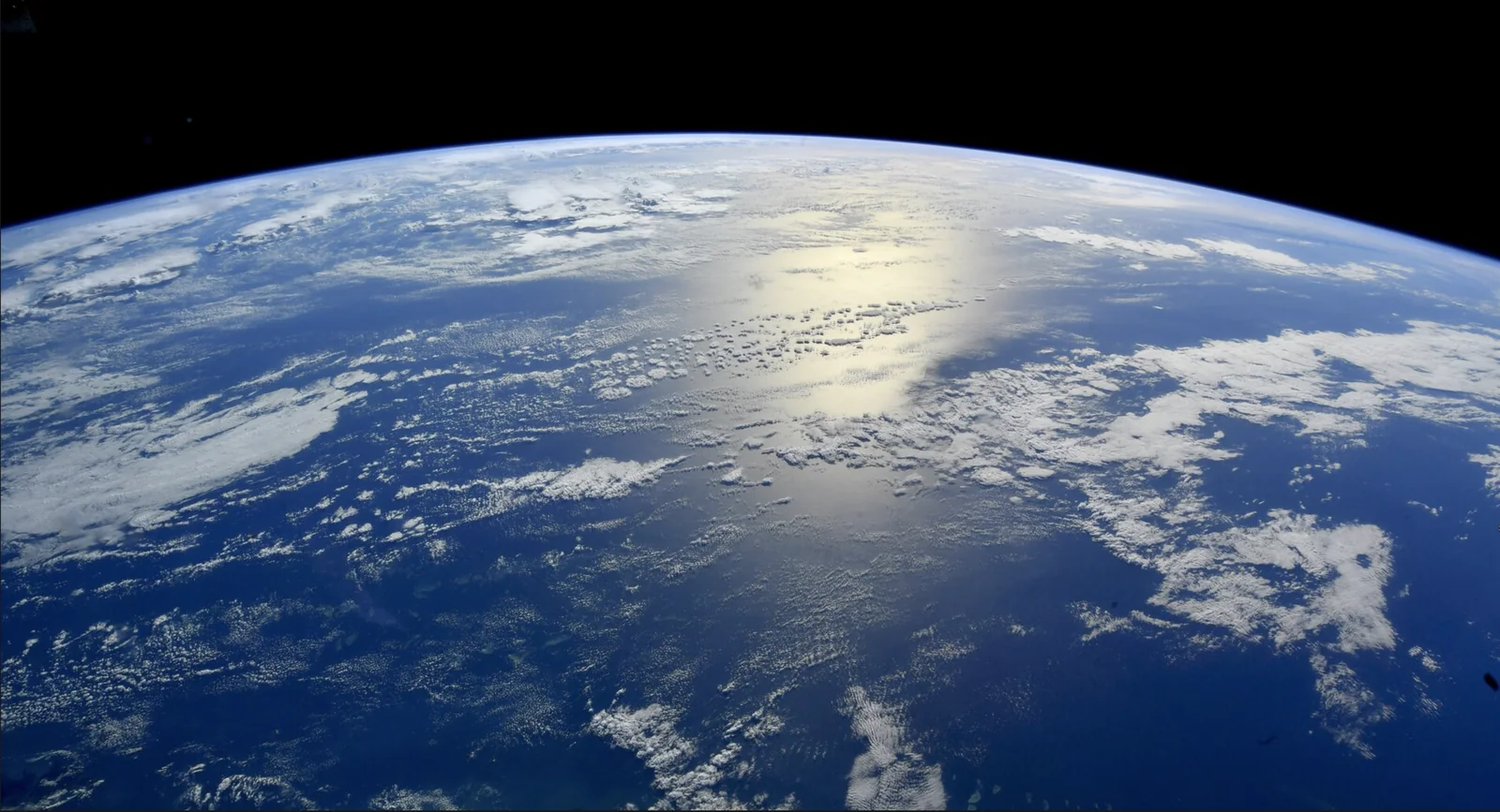 The curve of the Earth seen from orbit, with sunlight reflecting off the Pacific Ocean beneath broken cloud.